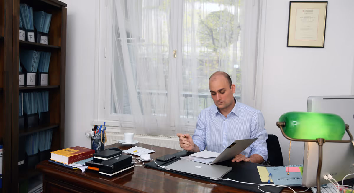 Man in a light blue shirt sitting at a desk in an office, reviewing documents with a laptop and desk lamp nearby.