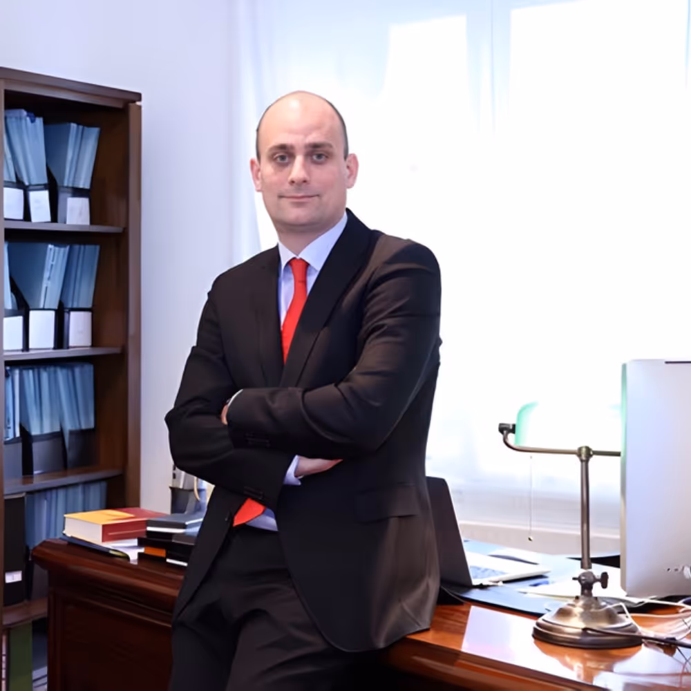 Man in a black suit and red tie leaning on a wooden desk in a bright office with books and files in the background.